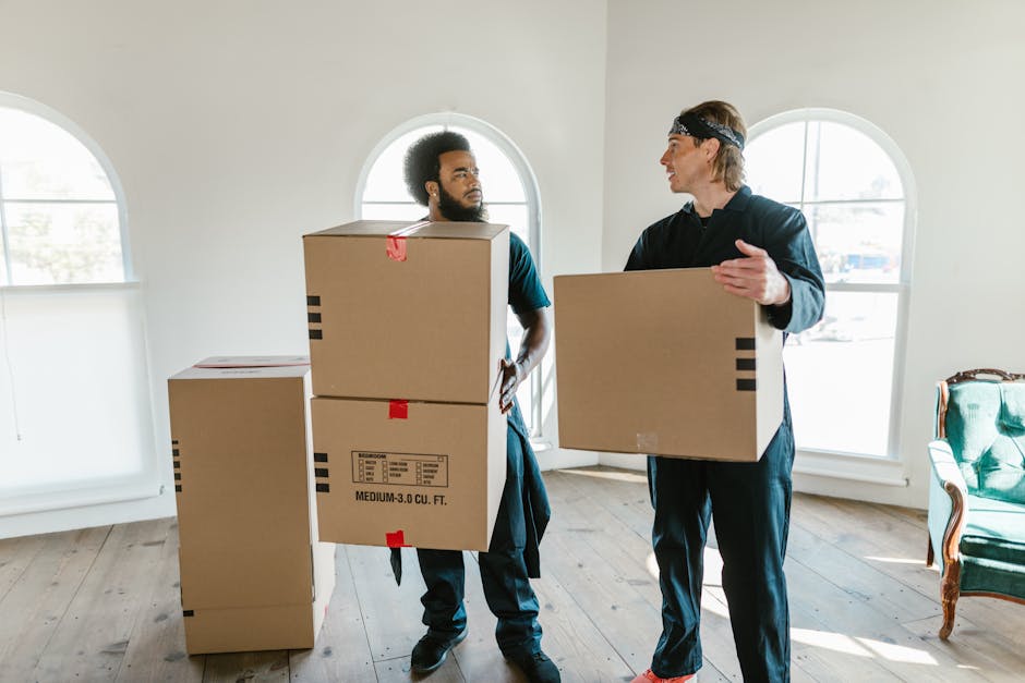 A man and woman standing inside a corridor beside a doorway, both holding cardboard boxes for home relocation; the man is holding a large box with red and black markings, wearing a maroon t-shirt and