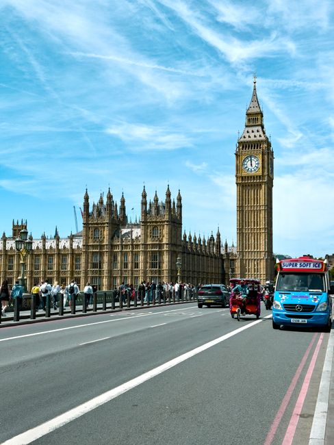 A busy street scene in Westminster with a view of Big Ben clock tower and the Houses of Parliament in the background under a partly cloudy sky. In the foreground, a white moving van is parked on the s