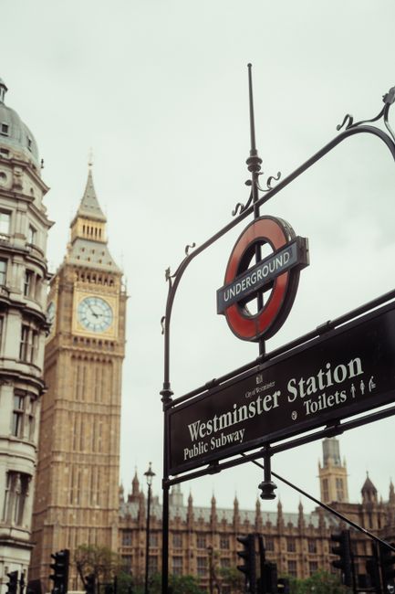 A London street scene outside Westminster station features a black underground signage sign with white lettering indicating 'Westminster Station' and additional directions for toilets and public subway access. Behind the sign, there is a view of the iconic Elizabeth Tower, commonly known as Big Ben, with its clock face visible, set against an overcast sky. To the left, partially visible, are historic stone buildings with detailed architectural features. In the foreground, there is a pavement area where a professional removal process may be occurring, involving the loading or unloading of furniture or moving boxes, although no specific personnel or equipment are visible. This scene underscores the busy and historic environment of Westminster, close to key transport and landmark sites, and subtly reflects themes of home relocation or furniture transport near Westminster underground station, as handled by Westminster Man and Van.